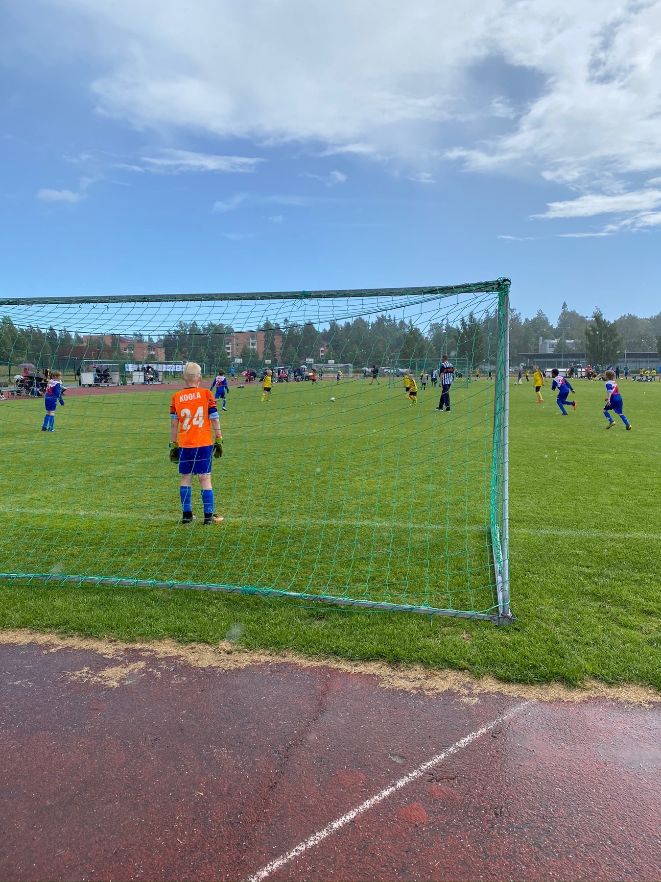 boy standing as a goal keeper in orange shirt, game going on further up field