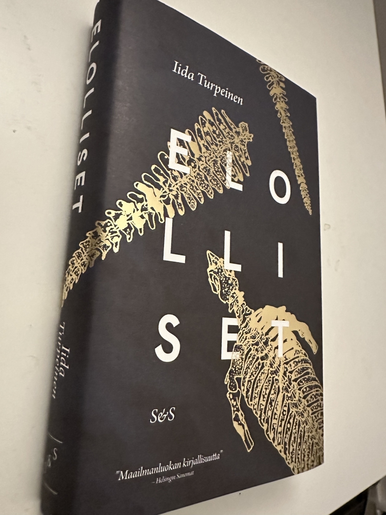 Hard book cover. It has the name of the author Iida Turpeinen and the book title Elolliset. On dark background there are two golden drawings of a skeleton of a Steller's sea cow