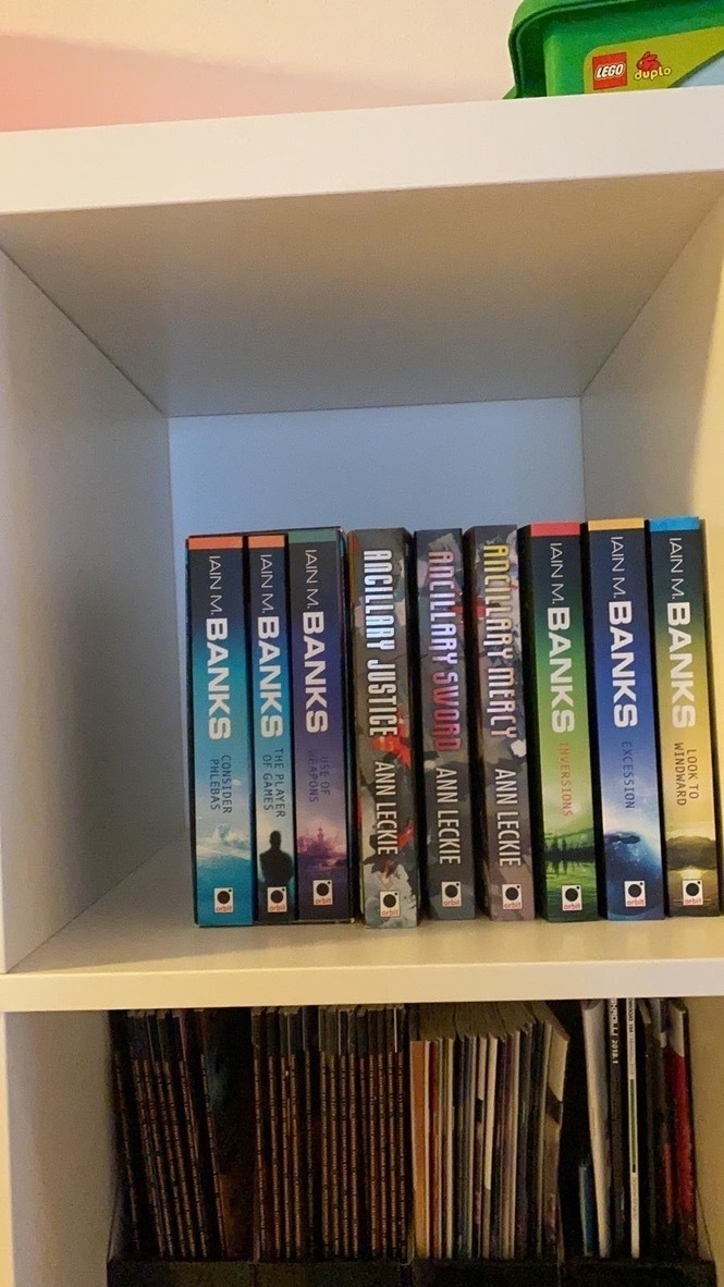 bookshelf with a set of Iain M. Banks books and Ann Leckie books. Magazines on lower shelf