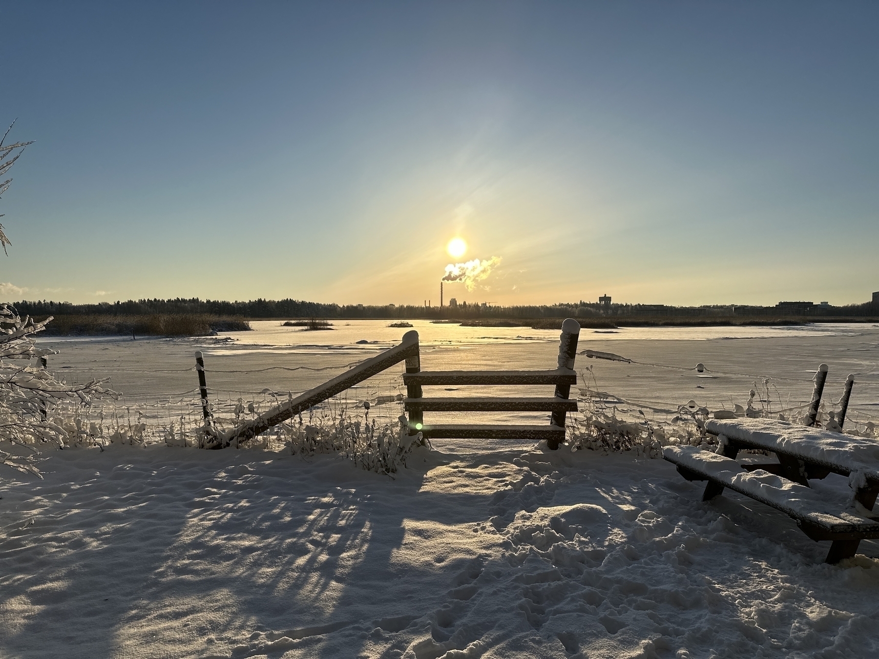 Midwinter sun seen over a field. Gate and fence shown in the front. A tall chimney seen in the distance, smoke billowing peacefully, giving sun a wintery beard